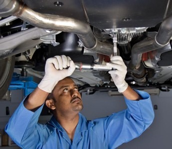 A man in blue overalls repairs a car, focused on the engine under the hood in a garage setting.